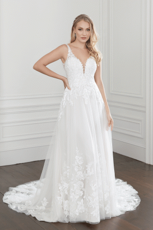 A woman stands posing in a room with white walls, wearing the Justin Alexander 44368 - Willow bridal gown. The white, sleeveless wedding dress features a deep V-neckline and intricate lace detailing. Its fitted bodice flows into a voluminous skirt with a long train. She has long, wavy blonde hair.