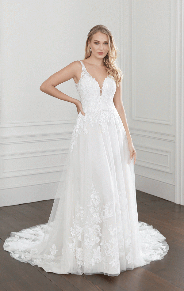 A woman stands posing in a room with white walls, wearing the Justin Alexander 44368 - Willow bridal gown. The white, sleeveless wedding dress features a deep V-neckline and intricate lace detailing. Its fitted bodice flows into a voluminous skirt with a long train. She has long, wavy blonde hair.