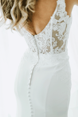 A woman wearing a white Stella York 7568 - Miami crepe and lace fitted wedding dress at Blessings Of Brighton, with a deep V-back and intricate floral lacework on the bodice. The back of the dress is fastened with covered buttons that run down to the skirt. Only the back and part of the torso are visible in the image.