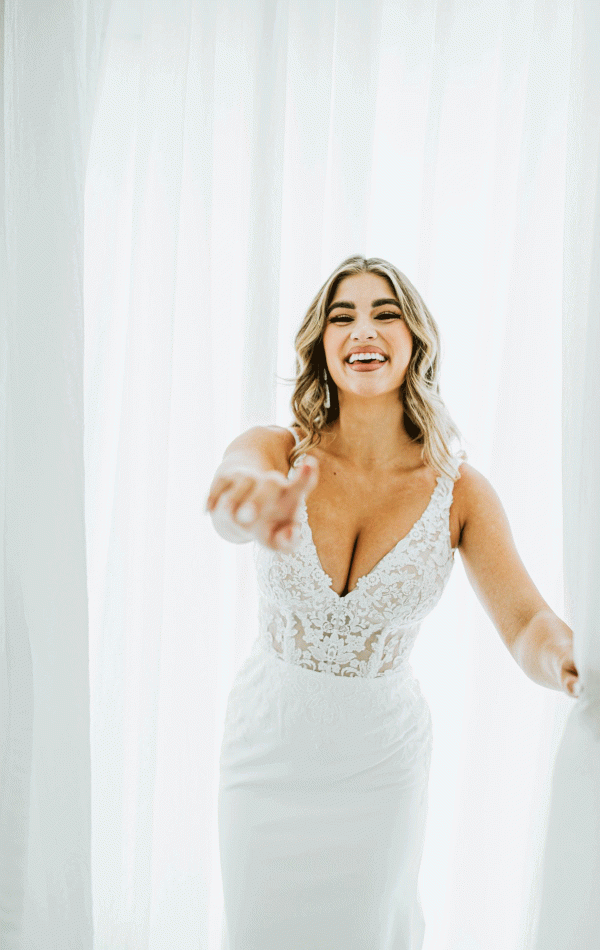 A woman in a white Miami (Style 7568) crepe and lace wedding dress by Stella York at Blessings Of Brighton stands in front of sheer white curtains, smiling widely and reaching forward with her left hand. The fitted bodice features intricate lace detailing and a deep V-neckline. Her hair is styled in loose waves.