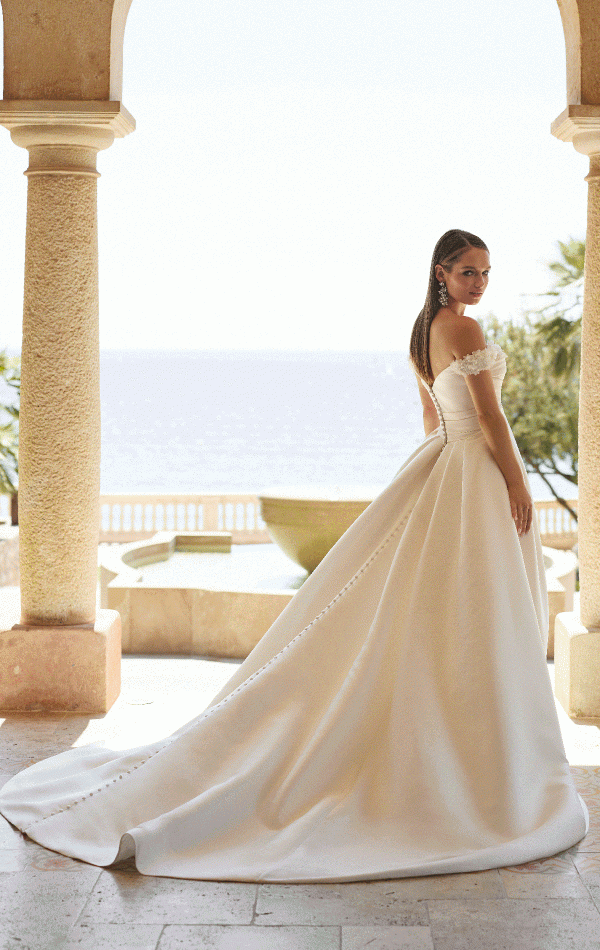 A bride stands in a scenic, sunlit archway overlooking a body of water. Dressed in the exquisite Ronald Joyce 18768 - Yoneko off-the-shoulder wedding gown with a long, flowing train, she exudes elegance amidst the setting's columns and fountain. In the distance, palm trees sway gently in the breeze.