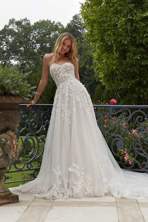 A woman in a strapless, white lace wedding gown from the 2625 - Paulette collection stands outside on a sunny day, leaning against an ornate metal railing. She is surrounded by lush greenery and vibrant flowers, with trees in the background. With shoulder-length blonde hair, she smiles at the camera.