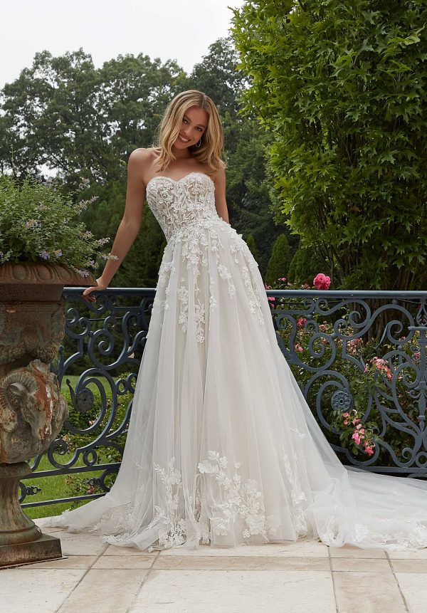 A woman in a strapless, white lace wedding gown from the 2625 - Paulette collection stands outside on a sunny day, leaning against an ornate metal railing. She is surrounded by lush greenery and vibrant flowers, with trees in the background. With shoulder-length blonde hair, she smiles at the camera.