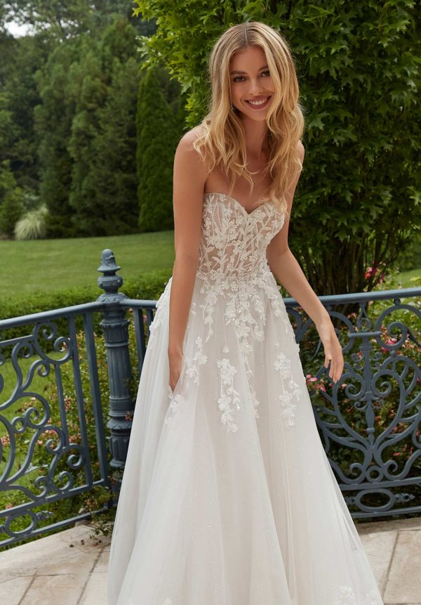 A woman, Paulette, with a radiant smile and long, wavy blonde hair is elegantly dressed in the 2625 - Paulette wedding gown. This stunning white, strapless lace dress features intricate floral appliqués. She is standing outdoors on a stone patio, surrounded by greenery and an ornate blue metal railing in the background.