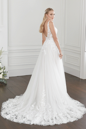 A bride stands in a white room with wood floors, wearing an elegant Justin Alexander 44368 - Willow bridal gown with lace details on the bodice and a flowing train. Her long, blonde hair is styled in loose waves, adorned with small floral accessories. She looks over her shoulder, smiling.