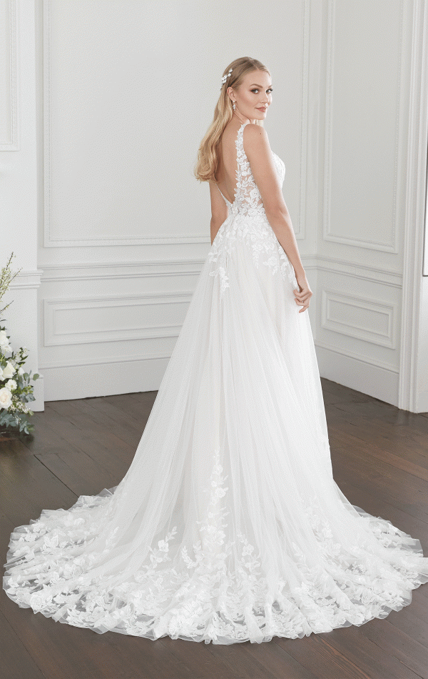 A bride stands in a white room with wood floors, wearing an elegant Justin Alexander 44368 - Willow bridal gown with lace details on the bodice and a flowing train. Her long, blonde hair is styled in loose waves, adorned with small floral accessories. She looks over her shoulder, smiling.