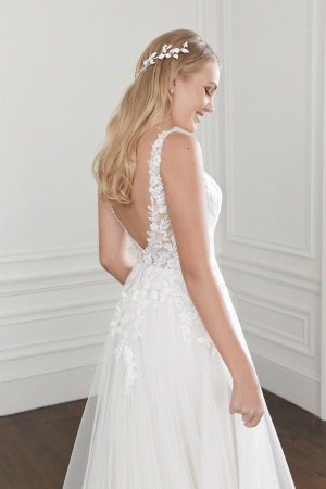 A bride with long blonde hair stands in a white room, wearing the Justin Alexander 44368 - Willow wedding gown, featuring delicate lace accents and an open back. With a delicate Willow floral hairpiece adorning her hair, she looks over her shoulder with a slight smile.