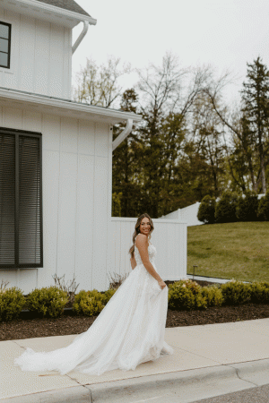 A bride in a Stella York 7790 - Evie wedding dress, featuring light tulle and 3D floral detail, stands on a sidewalk in front of a modern white building. She smiles while gently lifting her dress with one hand. The background includes trees and a grassy area.