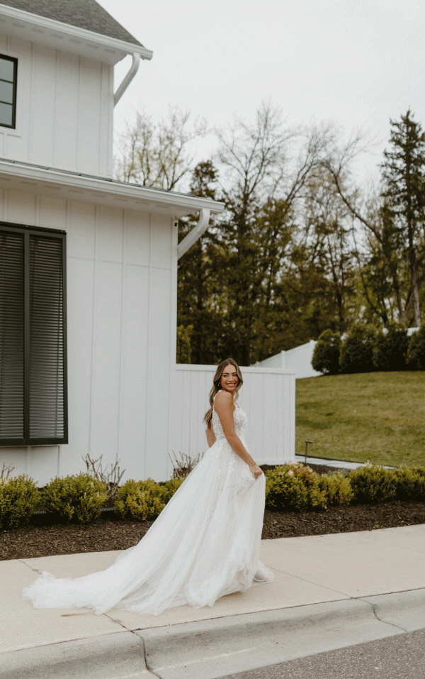 A bride in a Stella York 7790 - Evie wedding dress, featuring light tulle and 3D floral detail, stands on a sidewalk in front of a modern white building. She smiles while gently lifting her dress with one hand. The background includes trees and a grassy area.