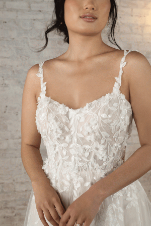 A woman is wearing the Stella York 7790 - Evie, a white, light Tulle A-line wedding dress adorned with 3D floral details and thin straps. Standing against a light-textured brick wall, her dark hair is styled in a loose updo, and her hands are gently clasped at her waist. She has a calm, contemplative expression.