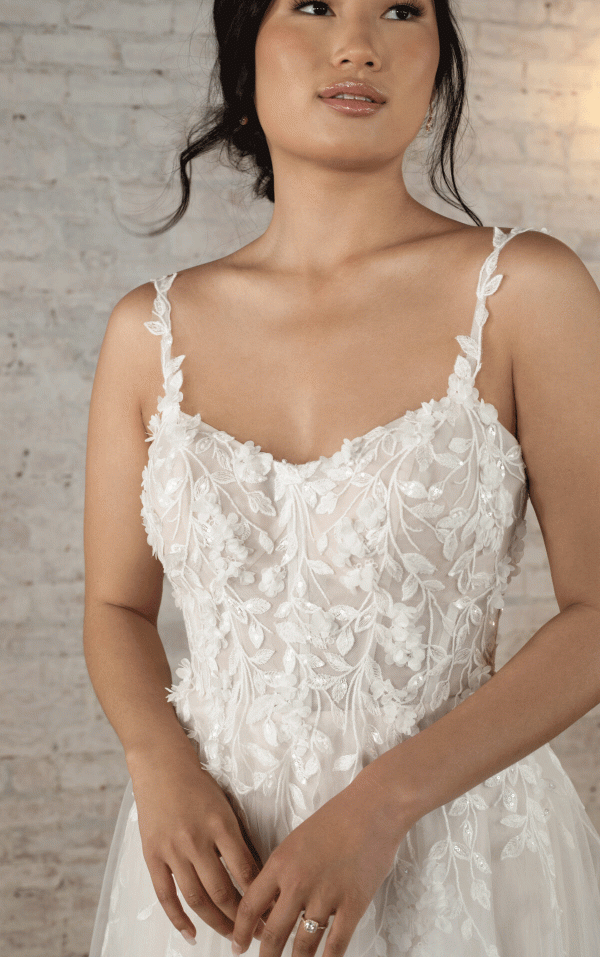 A woman is wearing the Stella York 7790 - Evie, a white, light Tulle A-line wedding dress adorned with 3D floral details and thin straps. Standing against a light-textured brick wall, her dark hair is styled in a loose updo, and her hands are gently clasped at her waist. She has a calm, contemplative expression.