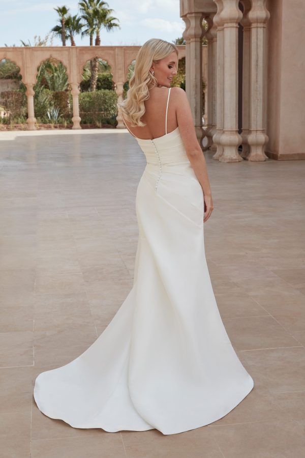 A woman with long blonde hair stands with her back to the camera, wearing a strapless white Justin Alexander 44424 - London-A wedding dress that features a fitted bodice and flowing skirt. She is outside on a stone patio, surrounded by arches, greenery, and palm trees.