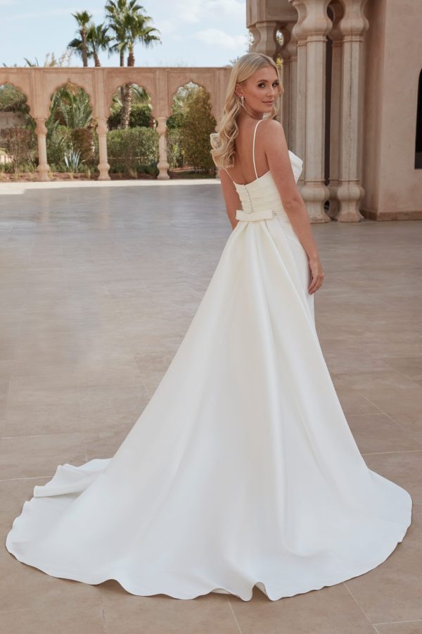 A bride in a stunning Justin Alexander 44424 - London-A white wedding gown with a long train stands outdoors in a sunny courtyard. The gown features a strapless bodice with a wide waistband tied into a bow at the back. She is looking over her shoulder, with stone columns and greenery in the background.