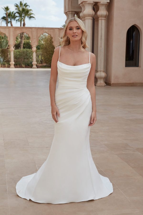 A woman stands in an outdoor architectural setting wearing the Justin Alexander 44424 - London-A white satin wedding gown, which features thin straps, a draped cowl neckline, and a fitted silhouette that flares slightly at the bottom. She has blonde hair styled in loose waves and an expression of calm confidence.