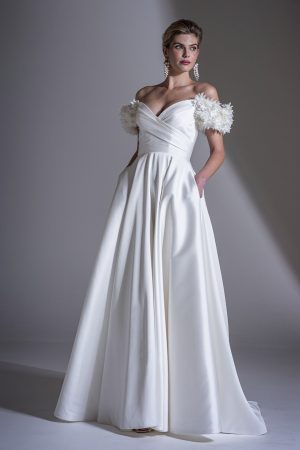 In a studio with a neutral backdrop, a woman is elegantly attired in the Freda Bennet - Romily Mikado wedding dress, featuring an off-the-shoulder neckline, floral appliqué sleeves, and convenient pockets. She complements her look with dangling earrings and an updo hairstyle that accentuates her stunning ensemble.