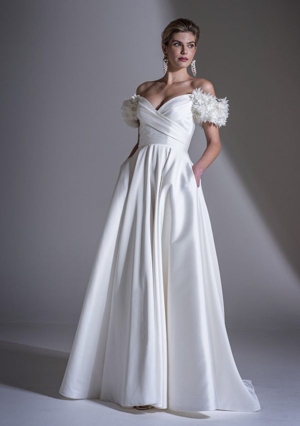 In a studio with a neutral backdrop, a woman is elegantly attired in the Freda Bennet - Romily Mikado wedding dress, featuring an off-the-shoulder neckline, floral appliqué sleeves, and convenient pockets. She complements her look with dangling earrings and an updo hairstyle that accentuates her stunning ensemble.