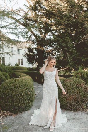 A bride in a Stella York 7941 - Leonie lace wedding dress with a thigh-high slit and scoop neckline stands in an outdoor garden at Blessings of Brighton, surrounded by manicured bushes and trees. She is looking down and touching her hair, with a tranquil expression on her face.