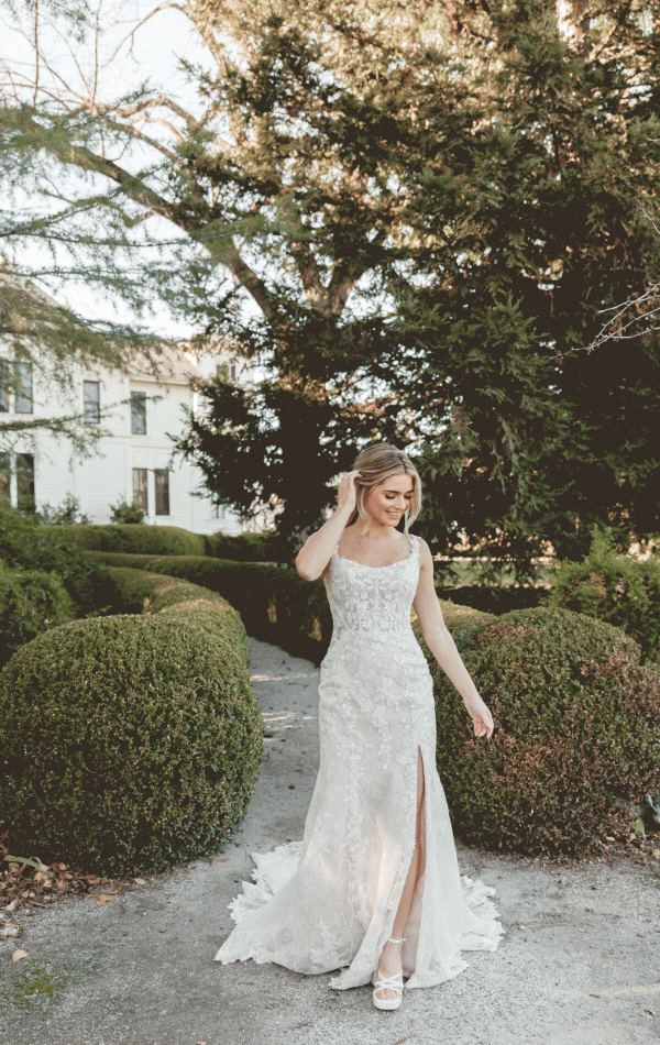 A bride in a Stella York 7941 - Leonie lace wedding dress with a thigh-high slit and scoop neckline stands in an outdoor garden at Blessings of Brighton, surrounded by manicured bushes and trees. She is looking down and touching her hair, with a tranquil expression on her face.