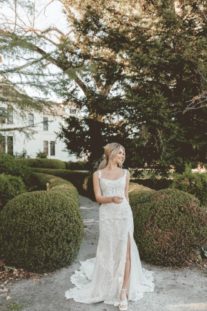 A bride in a Stella York 7941 - Leonie lace wedding dress with a scoop neckline and lace train stands on a garden path at Blessings of Brighton, surrounded by neatly trimmed bushes and trees. She has blonde hair styled in loose waves and is smiling while looking to her left, with a grand building visible in the background.