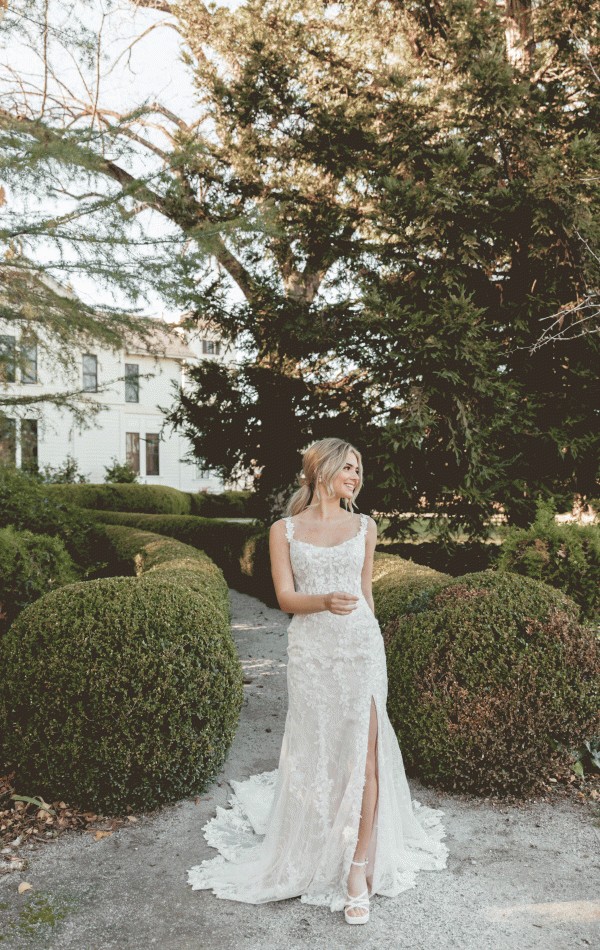 A bride in a Stella York 7941 - Leonie lace wedding dress with a scoop neckline and lace train stands on a garden path at Blessings of Brighton, surrounded by neatly trimmed bushes and trees. She has blonde hair styled in loose waves and is smiling while looking to her left, with a grand building visible in the background.