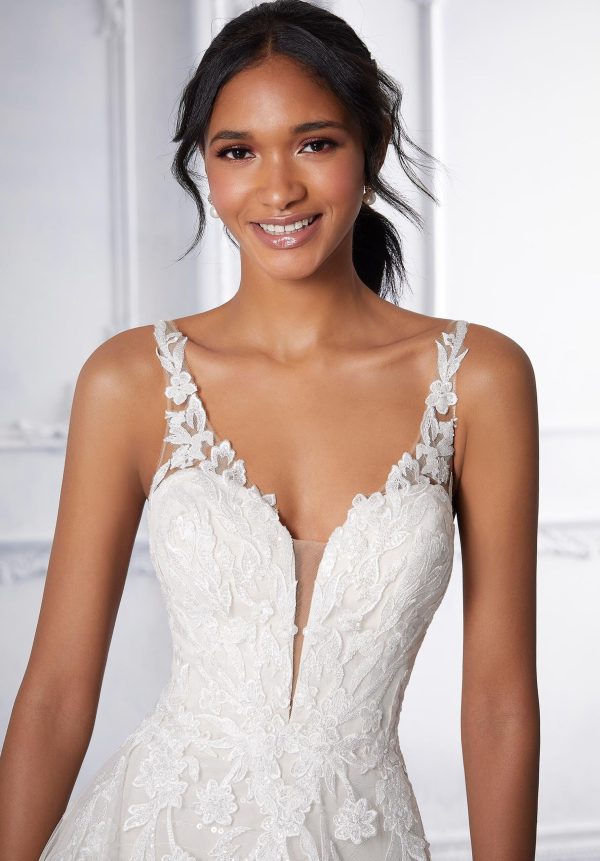 A smiling woman is wearing the Mori Lee 2378 - Calanthe, a sleeveless wedding gown adorned with intricate lace embroidery and floral details, featuring a deep V-neck. Her dark hair is tied back in a low ponytail as she stands against a light, softly blurred background. The design exudes elegance and sophistication.