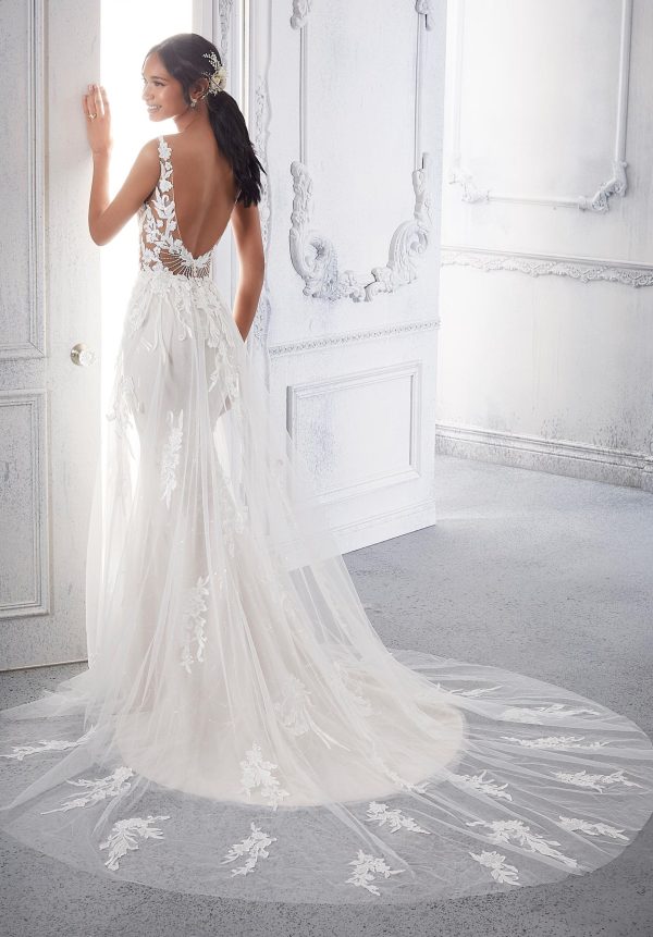 A woman stands by an open door wearing the stunning Mori Lee 2378 - Calanthe wedding dress, which features a low back and sleeveless design adorned with lace appliqués. Her long, dark hair is styled in a loose ponytail with a delicate hairpiece. The Calanthe dress boasts a long, sheer train with intricate lace details extending onto the floor.