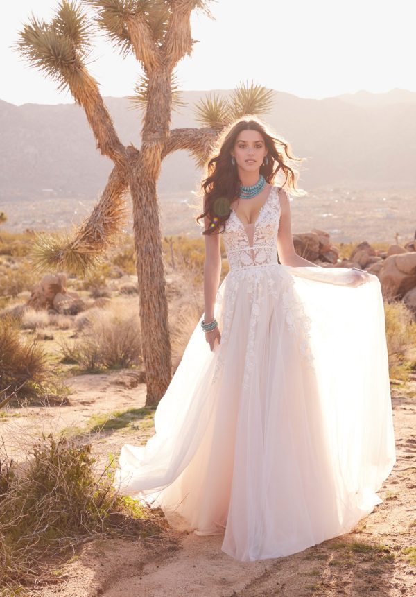 A woman in a flowing Mori Lee 2378 - Calanthe bridal gown with lace detailing stands in a desert landscape with mountains in the background. Her hair is loose, and she wears turquoise jewelry. She poses gracefully beside a Joshua tree under soft sunlight, evoking the elegance of Calanthe.