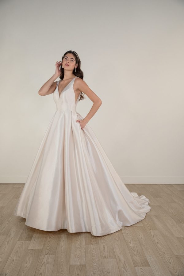 A woman stands in an elegant Stella York 7953 wedding dress with a deep V-neckline, sleeveless design, and a full skirt that gracefully grazes the floor. She poses with one hand on her waist and the other gently touching her hair against a simple indoor background with wooden flooring.