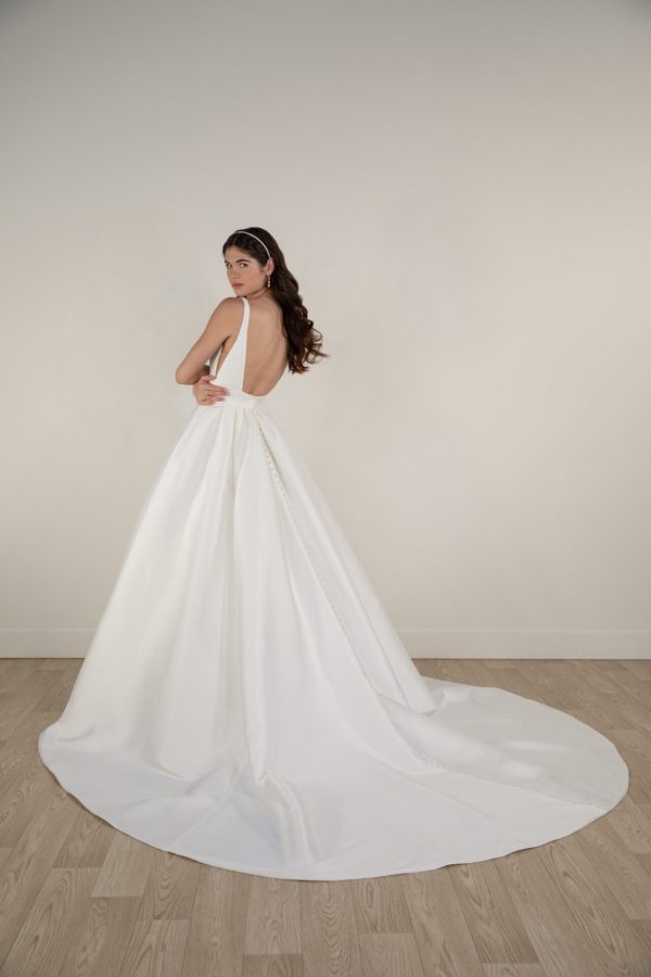 A woman stands on a wooden floor, facing left and looking back at the camera. She is wearing a stunning Stella York 7953 bridal gown with a long train that elegantly extends on the floor. Her dark hair is styled with a headband, and she poses with her hand on her waist, exuding timeless grace.