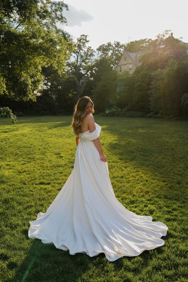 A woman wearing an elegant white off-the-shoulder Stella York 7644 - Kensington gown stands on a grassy field, bathed in sunlight. She is looking back over her shoulder, and her long, wavy hair cascades down her back. The background features lush trees and a building partially visible through the foliage.