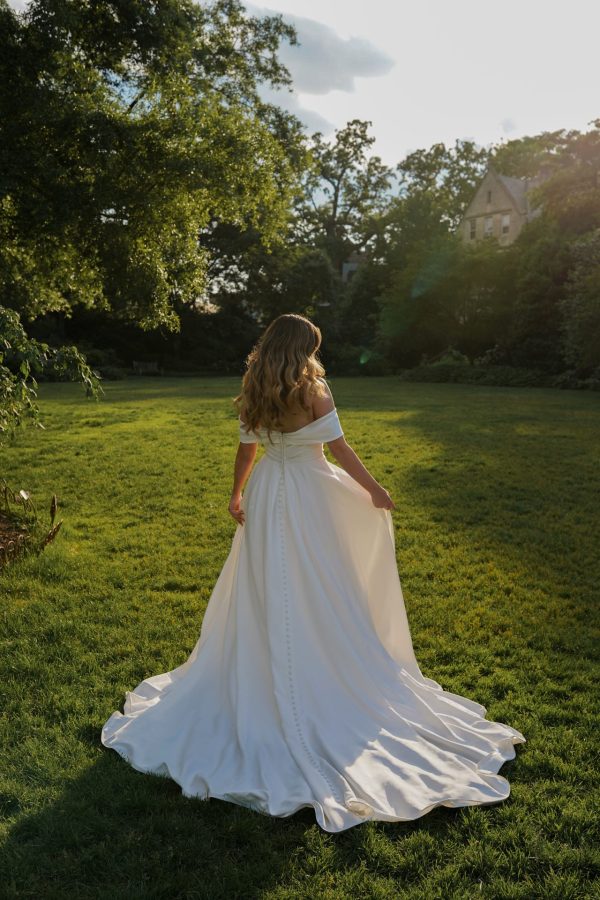 A woman with flowing blonde hair stands outdoors on a bright green lawn, wearing the Stella York 7644 - Kensington off-the-shoulder white wedding gown. She faces away from the camera, holding the edges of her dress, while large trees and a house are visible in the background.