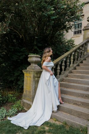 A woman in a Stella York 7644 - Kensington off-the-shoulder white gown ascends an outdoor stone staircase. Her long dress trails behind her, and she has long, wavy hair. She is wearing high-heeled shoes as she moves up the stairs beside a stone balustrade with lush greenery in the background.