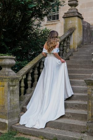 A woman in a long white Stella York 7644 - Kensington dress with a train walks up a stone staircase outside an old Kensington building. The staircase has moss-covered balustrades, and the surrounding greenery includes shrubs and trees, creating a lush, natural setting.