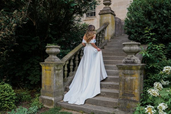 A woman in a long white Stella York 7644 - Kensington dress with a train walks up a stone staircase outside an old Kensington building. The staircase has moss-covered balustrades, and the surrounding greenery includes shrubs and trees, creating a lush, natural setting.