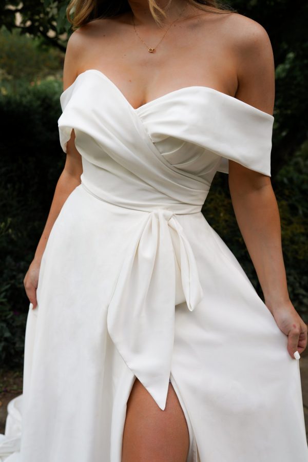 A woman in an elegant, off-the-shoulder Stella York 7644 - Kensington gown, featuring a wrap-style bodice and a thigh-high slit, stands outdoors surrounded by greenery. Her hair flows down beautifully as she holds the skirt of her dress with one hand.