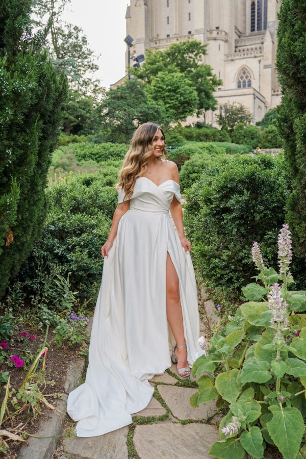 A woman in a long, white off-the-shoulder Stella York 7644 - Kensington dress stands on a stone path surrounded by lush greenery and plants. She has long, wavy hair and is smiling while holding the sides of her dress. The background features a tall, ornate building partially visible through the Kensington trees.