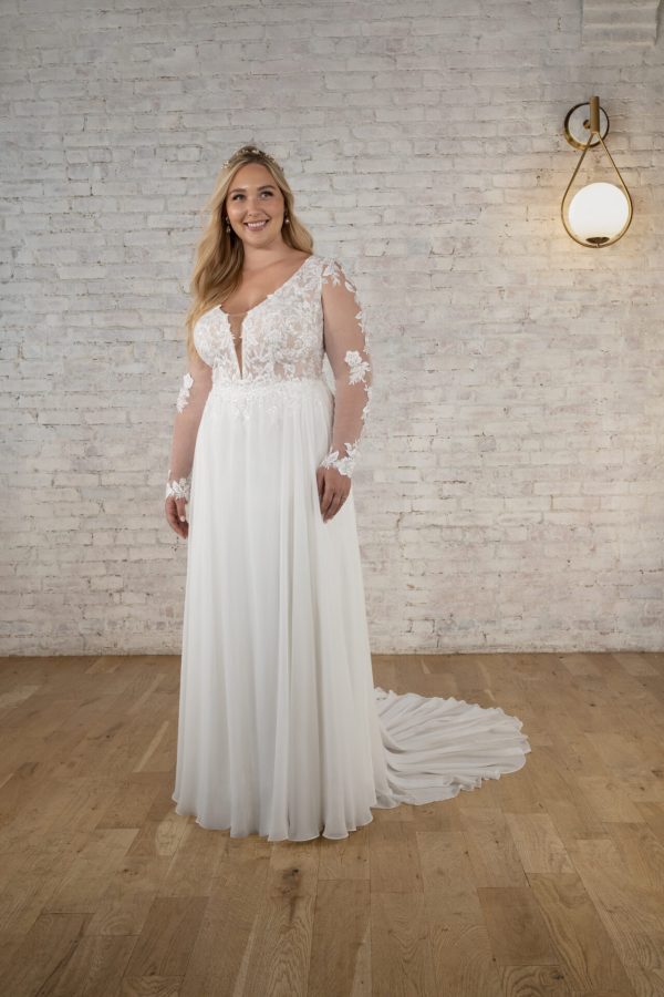 A woman stands smiling in a white Stella York 7699 - Ghalia wedding dress, which features long, sheer lace sleeves and a flowing skirt. She has long blonde hair and is wearing a tiara. The background showcases a white brick wall and wooden floor, complemented by a modern light fixture on the wall.