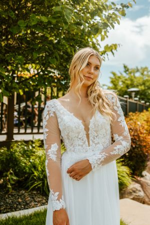 A blonde woman in a white, long-sleeved, lace Stella York 7699 - Ghalia wedding dress stands outdoors in a garden. She has loose waves in her hair and is looking off to the side with a serene expression. Greenery and a fence are visible in the background.