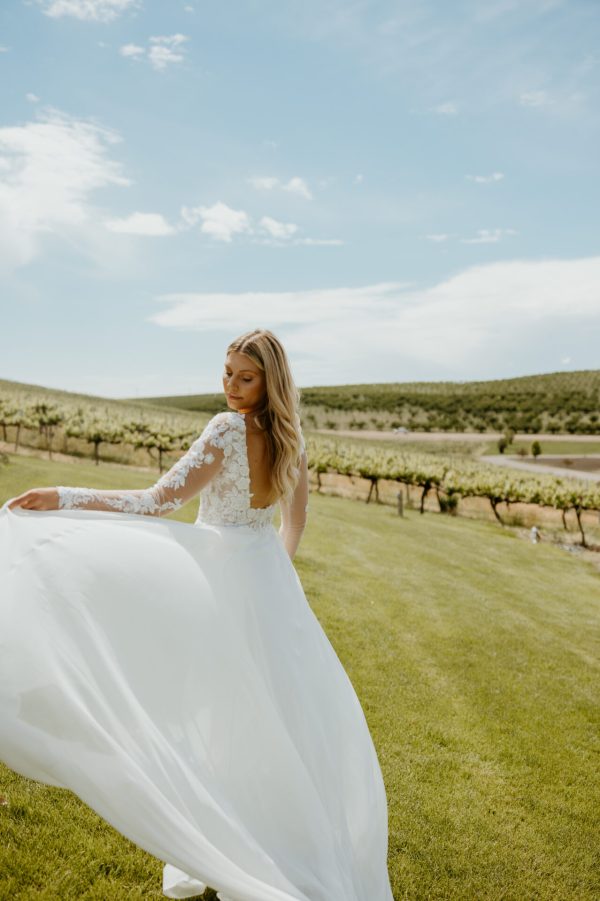 A bride with long blonde hair stands outdoors on a green lawn with vineyards in the background. She is wearing the stunning Stella York 7699 - Ghalia wedding dress, featuring lace long sleeves and a low back, holding out the skirt of her dress as it swirls around her. The clear, blue sky adds to the perfect moment.