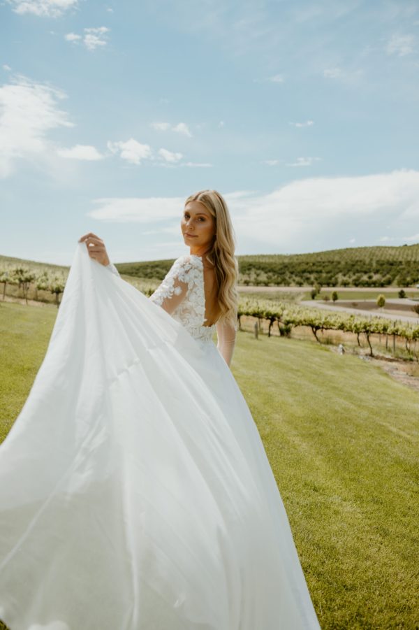 A bride with long blonde hair is standing outdoors on a grassy field. She is wearing a white Stella York 7699 - Ghalia wedding dress with lace sleeves and holding her dress up slightly as she turns to face the camera. Behind her, there is a vineyard under a partly cloudy sky.