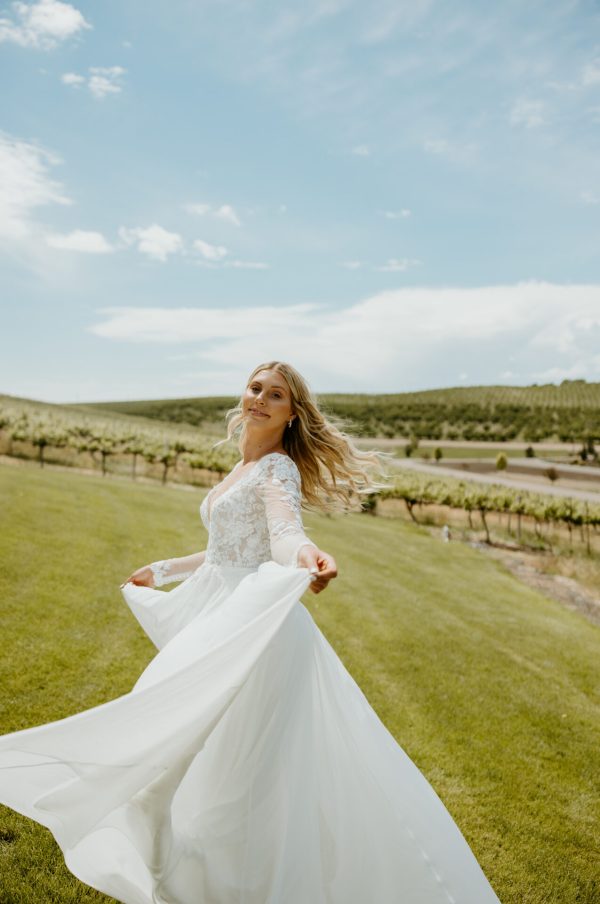 A woman in the Stella York 7699 - Ghalia wedding dress with lace sleeves is twirling on a grassy field. She is looking over her shoulder, with her long, wavy hair flowing behind her. In the background, rows of grapevines stretch under a blue sky with scattered clouds.