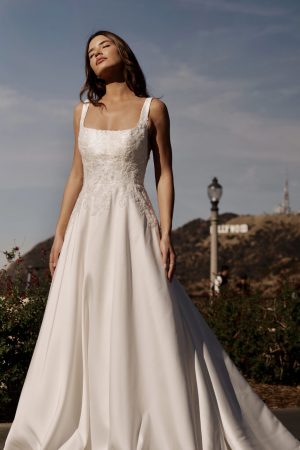 A woman stands outdoors donning the exquisite Stella York 7897 - Serene wedding dress, featuring intricate lace details on the bodice and a flowing skirt. She gazes upwards serenely, eyes closed and hands relaxed by her sides. A lamppost and a hillside with the Hollywood sign grace the background.