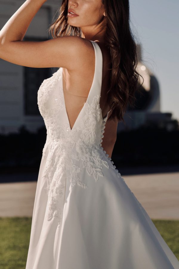 A woman wearing the stunning Stella York 7897 - Serene wedding dress with intricate lace detailing on the bodice and a flowing skirt stands outdoors on a sunny day, with her left hand raised near her head. The pose showcases the dress's sleeveless design and deep V-cut on the side.