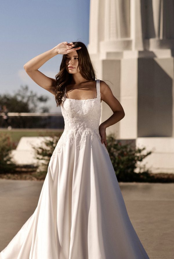 A serene woman in a stunning Stella York 7897 - Serene wedding dress stands outdoors with one hand shielding her eyes from the sun. The dress features intricate lace details on the bodice and flows into a long, elegant skirt. A tall, white stone structure is visible in the background.