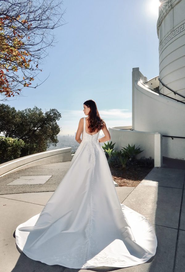 A serene bride stands outdoors with her back facing the camera, wearing the Stella York 7897 - Serene wedding dress, showcasing its long and flowing train. Positioned near a white building with curved architectural details, the sun casts a bright light, creating a clear shadow of her elegant gown.