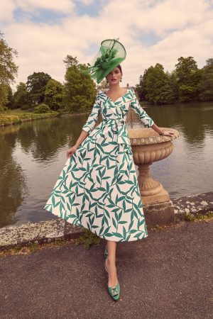 A woman, clad in the Veni Infantino 992266 white and green leaf-patterned dress, stands beside a stone railing near a lake. She wears a large green hat with a veil. Her head is tilted slightly, one hand on the railing, surrounded by trees and under a cloudy sky.