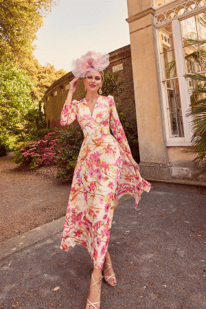 A person in a floral dress and an elegant pink headpiece stands gracefully, slightly tilting their head while holding the headpiece with one hand. They are outdoors near a historic building adorned with large windows, surrounded by greenery and blooming flowers—a scene perfectly captured by Invitations by Veni - 29662 for inspiration.