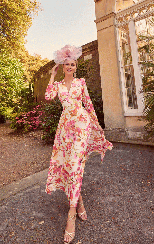 A person in a floral dress and an elegant pink headpiece stands gracefully, slightly tilting their head while holding the headpiece with one hand. They are outdoors near a historic building adorned with large windows, surrounded by greenery and blooming flowers—a scene perfectly captured by Invitations by Veni - 29662 for inspiration.