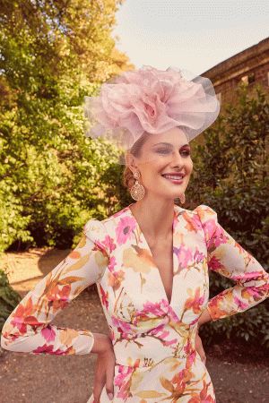 A woman stands outside, her smile beaming. She is dressed in a vibrant floral dress adorned with pink, yellow, and orange blossoms. Crowning her head is an elegant pink fascinator. Lush greenery and sunlit trees create the perfect backdrop for sending out Invitations by Veni - 29662 to celebrate the occasion.