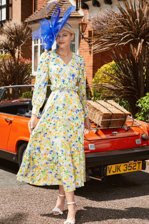 A person wearing a long, floral dress and a decorative blue hat stands on a street in front of an orange convertible car with a wicker basket on its back. The scene beautifully captures the essence of "Invitations by Veni - 29679," set against the backdrop of a brick house adorned with greenery and plants.